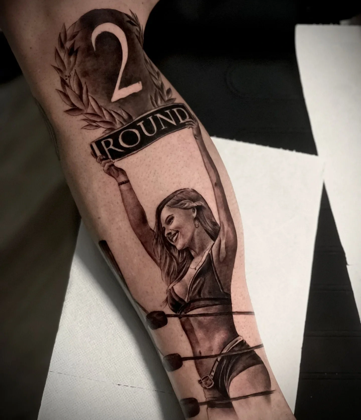 Round 2 boxing piece — laurel wreath with '2 ROUND' banner and woman holding placard between ring ropes